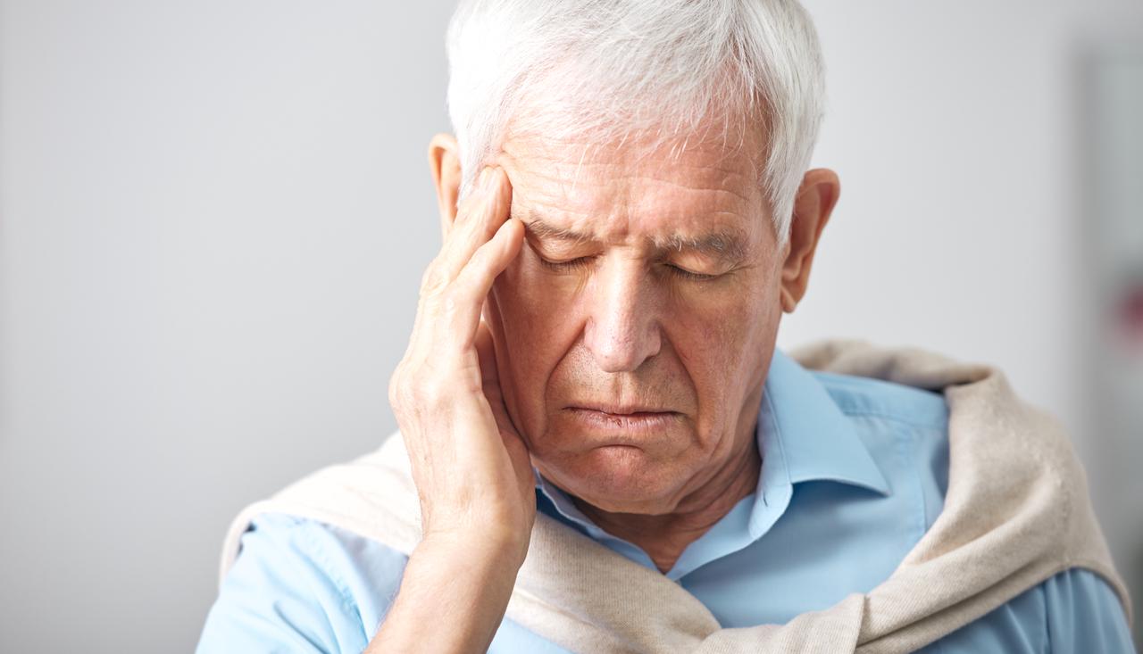 Elderly man presses his temple, eyes closed, suffering from headache against light neutral background