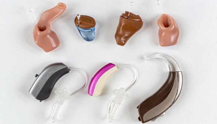Various styles of hearing aids displayed on white background, including custom in-ear IEM hearing aids and behind-the-ear BTE hearing aids with diverse colors and personalized ear molds for hearing loss assistance
