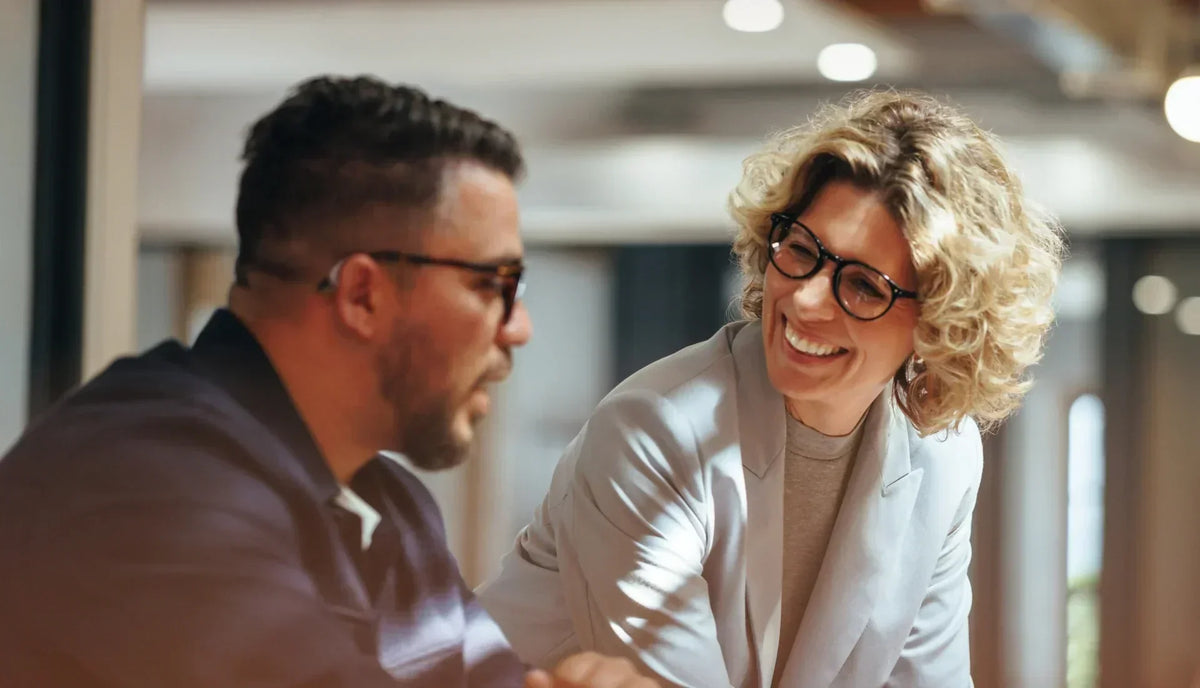 Businessman wearing ELEHEAR hearing aid talking with female colleague in modern office, smooth professional communication.