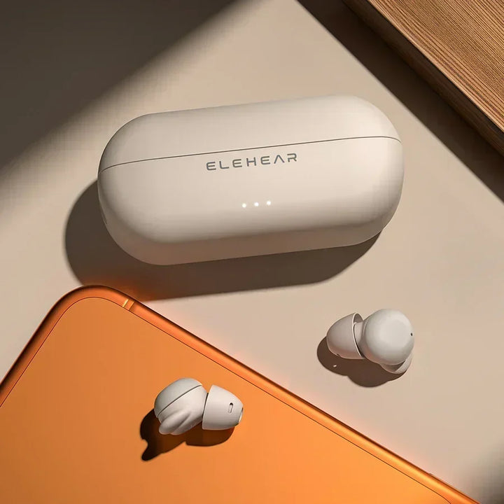 Beige ELEHEAR in-ear hearing aids with matching charging case, warm minimalist desktop scene with orange accent surface.