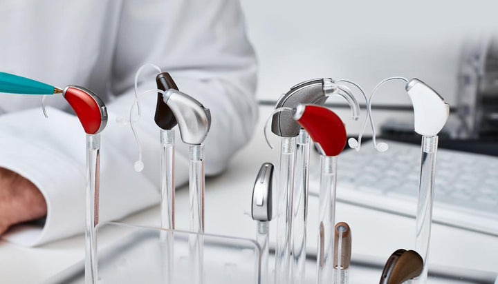 Various colorful behind-the-ear hearing aids displayed on clear acrylic stand in audiology clinic; audiologist uses tool to inspect hearing devices for hearing loss treatment and personalized fitting