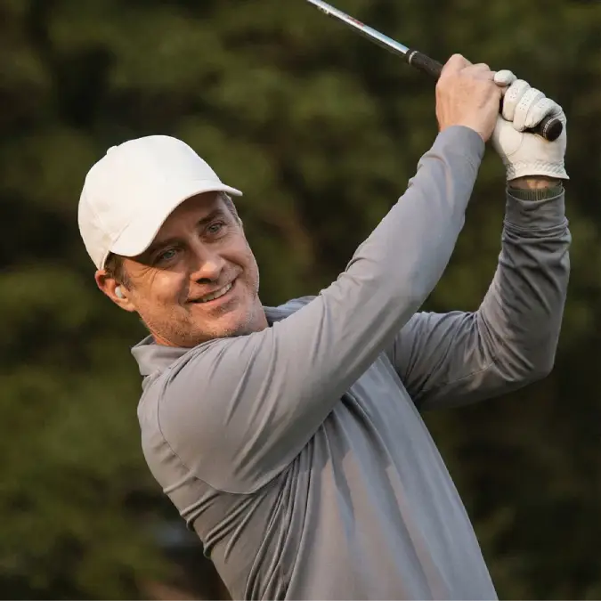 Smiling middle-aged man wearing ELEHEAR in-ear hearing aid swinging golf club on outdoor golf course with blurred green foliage background