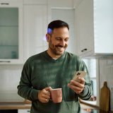 Smiling man wearing hearing aid with blue LED light while using smartphone and holding coffee mug in kitchen thumbnail