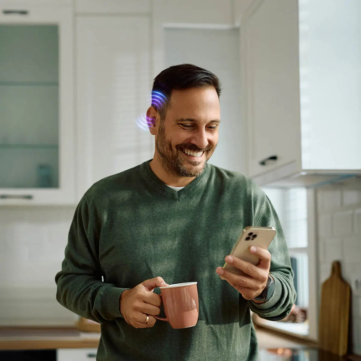 Smiling man wearing hearing aid with blue LED light while using smartphone and holding coffee mug in kitchen