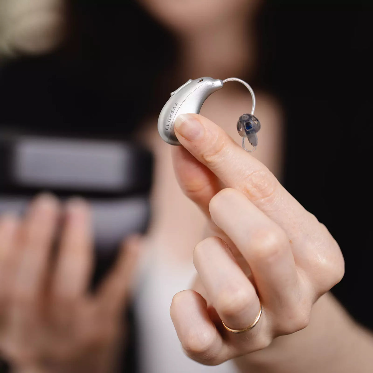 Hand holding silver ELEHEAR Beyond hearing aid with clear ear dome
